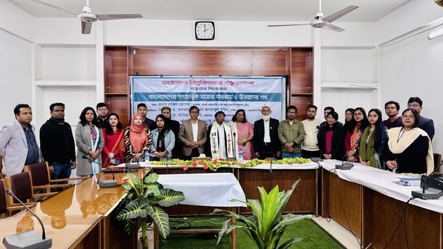 Nazim Hossain, wearing the cream ceremonial scarf, stands in the middle of a large group of men and women in a conference room, in front of the lecture banner.