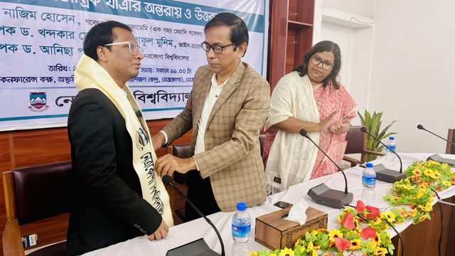 Nazim Hossain, in a dark suit and glasses, has a cream ceremonial scarf placed around his neck by a man in a checked jacket, while a woman in a white shawl and pink sari applauds beside a conference table. Behind them, a Bangla banner names the lecture and its speakers.